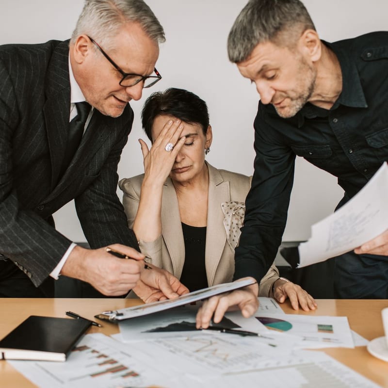 A manager with her head in her hand while two colleagues push papers at her.